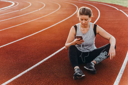 A young beautiful woman having rest with her smartphone sitting at a stadium running track.の写真素材