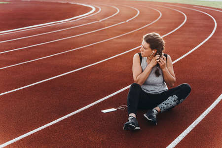 A young beautiful woman having rest with her smartphone sitting at a stadium running track.の写真素材