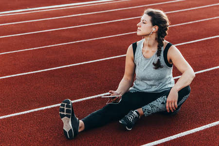 A young beautiful woman having rest with her smartphone sitting at a stadium running track.の写真素材