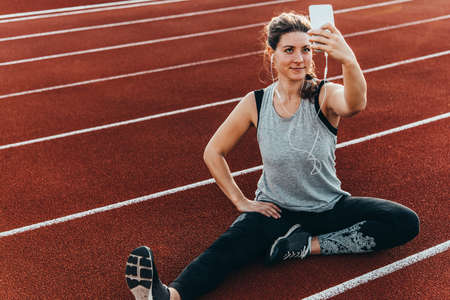 A young beautiful woman making selfie sitting at a stadium running track.の写真素材
