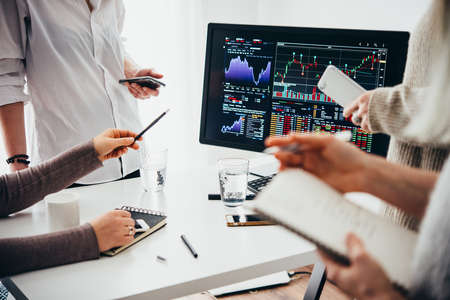 A group of colleagues discussing recent stock exchange trends pointing to the stock exchange screen showing trading graphicsの写真素材