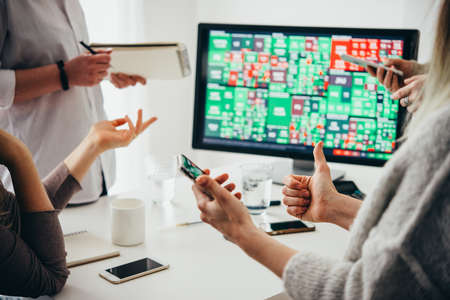 A group of colleagues discussing recent stock exchange trends pointing to the stock exchange screen showing trading graphicsの写真素材