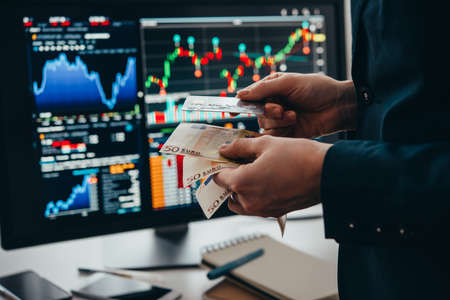 Hands holding european currency Euro and a credit card in front of the stock exchange screen showing trading graphics.の写真素材