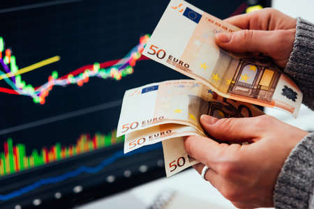 Hands holding european currency Euro in front of the stock exchange screen showing trading graphics.の写真素材