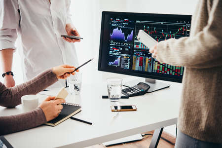 A group of colleagues discussing recent stock exchange trends pointing to the stock exchange screen showing trading graphicsの写真素材