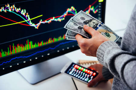 Hands holding US dollars currency in front of the stock exchange screen showing trading graphics.の写真素材