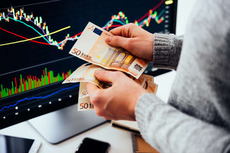 Hands holding european currency Euro in front of the stock exchange screen showing trading graphics.の写真素材