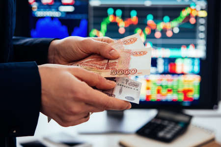 Hands holding russian currency roubles in front of the stock exchange screen showing trading graphics.の写真素材