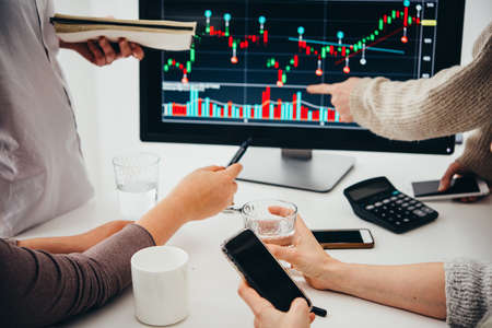 A group of colleagues discussing recent stock exchange trends pointing to the stock exchange screen showing trading graphicsの写真素材