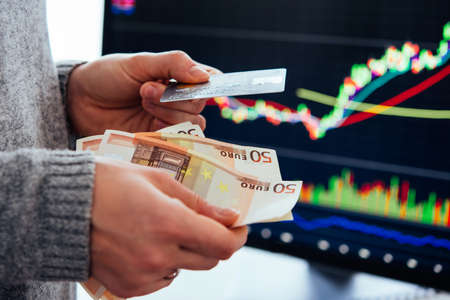 Hands holding european currency Euro and a credit card in front of the stock exchange screen showing trading graphics.の写真素材