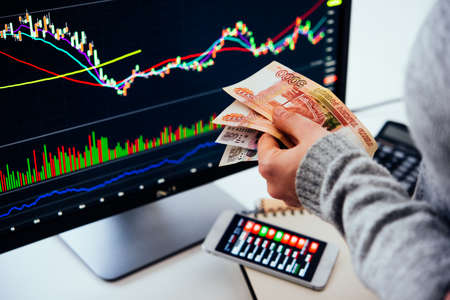 Hands holding russian currency roubles in front of the stock exchange screen showing trading graphics.の写真素材