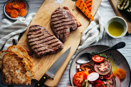 Steaks served on wooden board with tomato salad and grilled vegetables ready for dinner. Healthy home eating concept.の写真素材