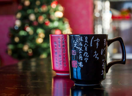 Two mugs red and black decorated with chinese calligraphy, lying on black chinese table likewise decorated with calligraphy signs with blurred christmas tree in backgroundのeditorial素材