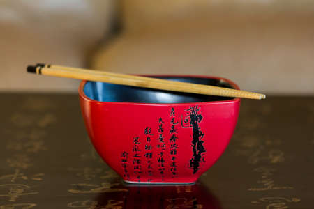 Red chinese rice bowl decorated with chinese calligraphy with chopsticks  on its top lying on the surface of black chinese table likewise decorated with calligraphyのeditorial素材