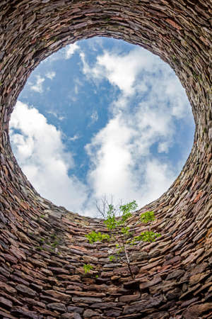 The inside of an old industrial chimney shaft photographed from the bottom - circular stone wall with tree growing from it and blue sky with white clouds in the opening in the centre, verticalの写真素材