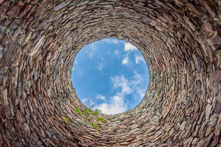 The inside of an old industrial chimmey shaft photographed from the bottom - circular stone wall with tree growing from it and blue sky with white clouds in the opening in the centre, horizontalの写真素材