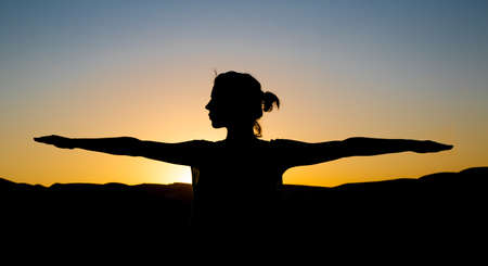 Silhouette of a woman against a beautiful sunset background. a Woman stretches her hands and doing yoga meditation exercise.の写真素材