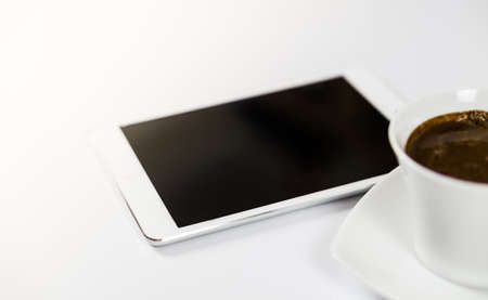 Computer tablet with empty screen next to coffee. The tablet is on the white table. View from the side with sunflare. Close-up.の写真素材