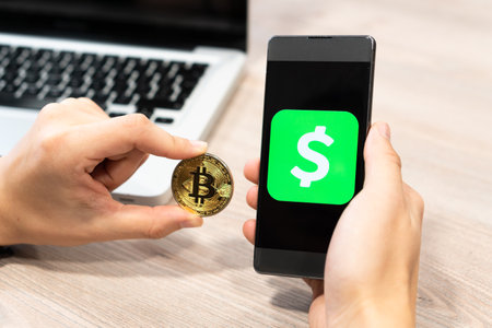 Cash App logo by Square inc displayed on smartphone held by human hand next to Bitcoin coin and computer laptop - Slovenia 13.02.2019のeditorial素材