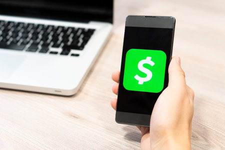Cash App logo by Square inc displayed on smartphone held by human hand next to computer laptop - Slovenia 13.02.2019のeditorial素材