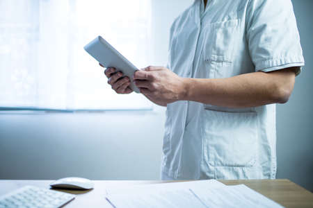 Doctor in the uniform using computer tablet next to his office desk in hospitalの写真素材