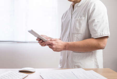 Doctor in the uniform using computer tablet next to his office desk in hospitalの写真素材
