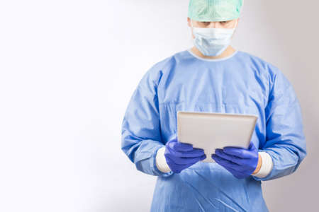 Surgeon doctor in sterile gloves preparing for operation using tablet computer. He is wearing surgical cap and blue gownの写真素材