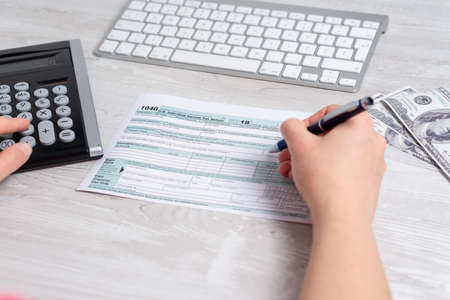Point of view photo of woman calculating and filling US tax form next to computer keyboard, dollar bills and tax form 1040. 15th April 2019 tax dayの写真素材