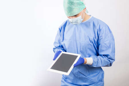 Surgeon doctor in sterile gloves preparing for operation using tablet computer. He is wearing surgical cap and blue gownの写真素材