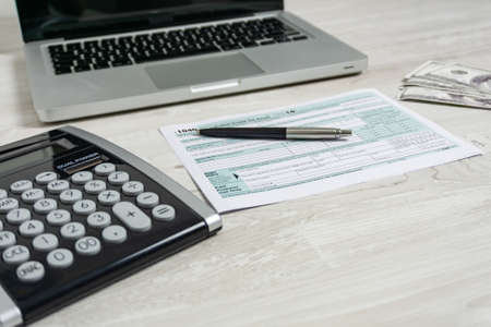 US tax form next to computer laptop, dollar bills, calculator and tax form 1040. tax form us business income office hand fill conceptの写真素材