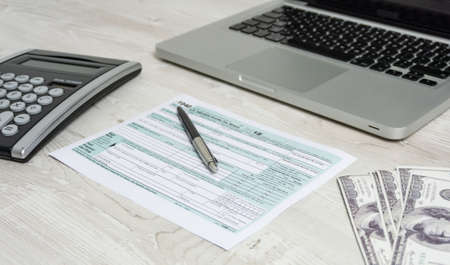 US tax form next to computer laptop, dollar bills, calculator and tax form 1040. tax form us business income office hand fill conceptの写真素材