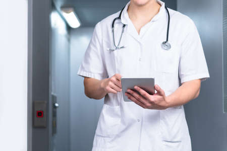 Bright photo of male doctor in uniform with stethoscope coming out of the elevator and using computer tablet in hospital. Modern medical conceptの写真素材