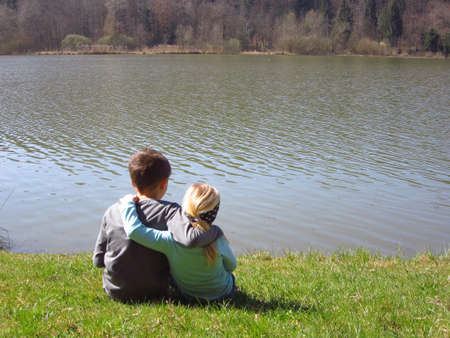 Little brother and sister by the lake hugging tight and beeing happy together in nature and outdoors. Girl and a boy sitting on a grassの写真素材