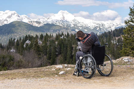 Disabled man on wheelchair using camera in nature, photographing beautiful mountainsの写真素材