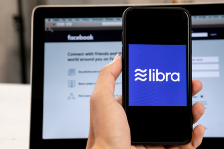 18 June 2019, Ljubljana Slovenia - Hand holding a smartphone with Libra logo on it, next to Facebook website opened on computer laptopのeditorial素材