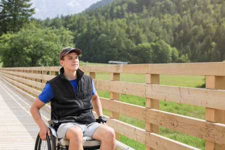 Disabled young man on a wheelchair on a wooden bridge path enjoying in nature looking at beautiful viewの写真素材
