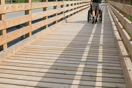 Assistant pushing wheelchair outside in nature on a wooden bridge pathの写真素材