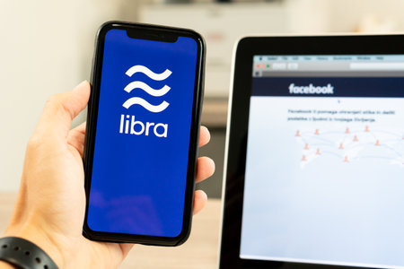 12 July 2019, Ljubljana Slovenia - Hand holding smartphone with Libra logo next to facebook webpage....