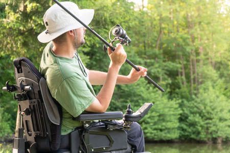 Happy man in a electric wheelchair throwing fishing pole at the beautiful pond in natue on a sunny dayの写真素材