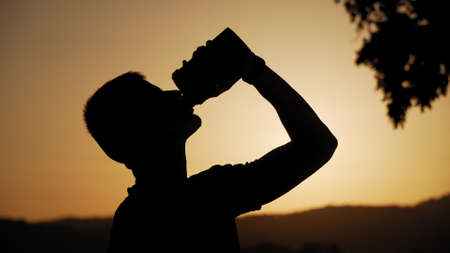 silhouette of fit young man drinking water with sun and nature behind it in sunset
