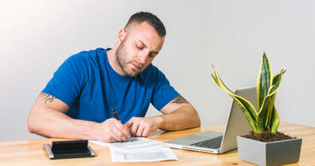 Man filling 1040 US tax form by hand with calculator and laptop.