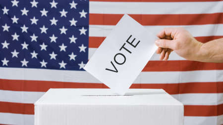Voting hand casting ballot in a box at flag of USA. American presidential elections.