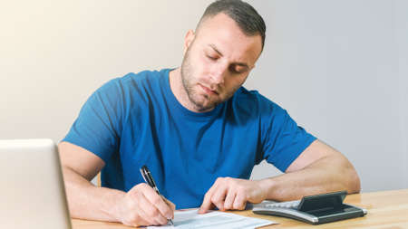 Young casual man filling 1040 tax form by hand with calculator and laptop.