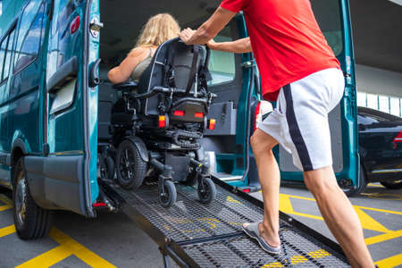 Driver assisting disabled person on wheelchair with transport using accessible van with ramp.