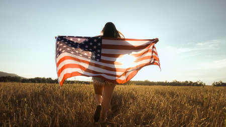 American teenager female in field of wheat holding USA flag in golden sunset with flare. 4th of July, Independence Day.の写真素材
