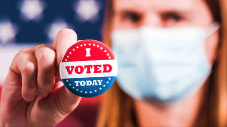 Woman with face mask showing I voted today button with american flag. Extreme close-upの写真素材