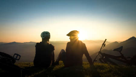 Two female cyclists sitting on the grass, talking and looking at mountains in the distance at the...