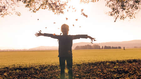 Smiling preschool girl, standing at the edge of the forest, holding a pile of dry leaves in hands, throwing it in the airの写真素材