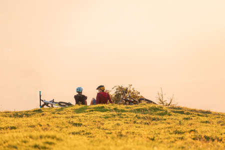 Two female cyclists sitting on the grass, talking and looking at mountains in the distance at the sunsetの写真素材