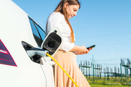 Woman plugging in the charger into her white electric car. Using a smartphone while waitingの写真素材